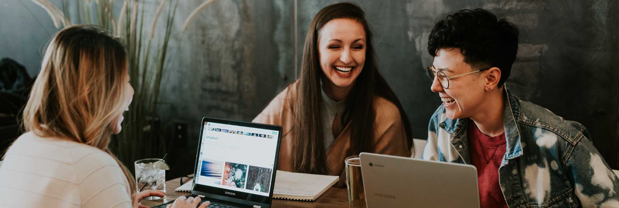 Three laughing women with laptops sitting around a table working.